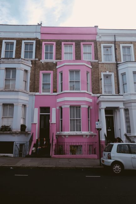 A row of Victorian terraced houses in Notting Hill, featuring a mix of white, pink, and blue facades with large sash windows and decorative bay windows. The buildings are constructed with brick, some 