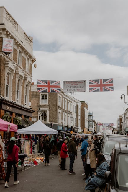 A busy street scene on Portobello Road in Notting Hill, featuring a row of multi-storey Victorian-style buildings with decorative facades. The street is filled with pedestrians and shoppers, some browsing market stalls that are covered with white and pink canopies, displaying various items such as clothing, accessories, and household goods. A grey vehicle is parked on the right side of the street, partially blocking the view of the sidewalk. Above, three Union Jack flags are strung across the street, along with a large banner that reads 'Portobello Road W11.' The scene is set during daytime with overcast skies, providing natural light that illuminates the street activity. This image captures the lively atmosphere typical of a home relocation or moving logistics context, as storage and transport services like those offered by Storage Notting Hill facilitate the packing, loading, and furniture transport associated with house removals in the Notting Hill area.