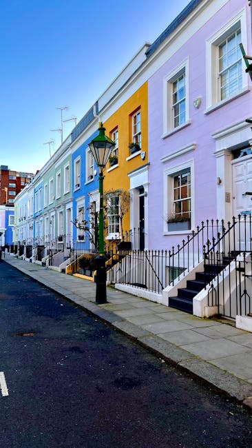 A row of three colourful terraced houses located on a residential street, with pastel pink, yellow, and blue facades. Each house has three visible white-framed sash windows, some with decorative mould